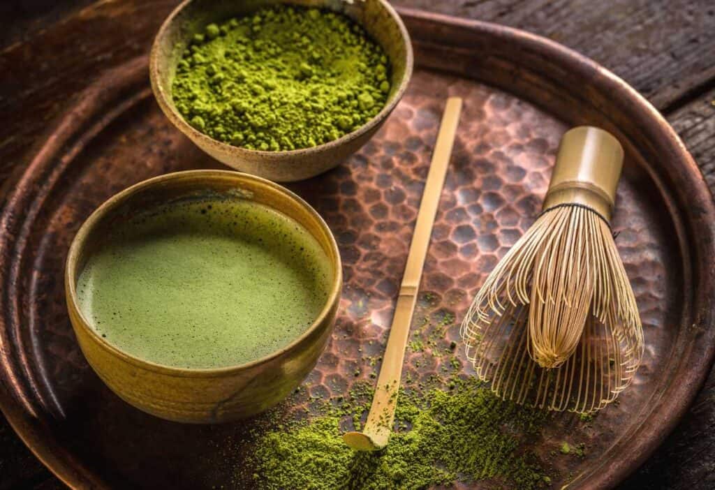 A bronze tray with a bowl of green matcha powder, a bamboo whisk and freshly made matcha tea.