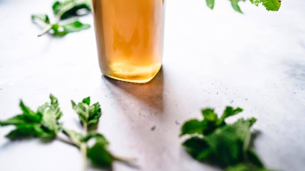 A clear glass bottle filled a light yellow syrup surrounded by fresh green mint leaves.