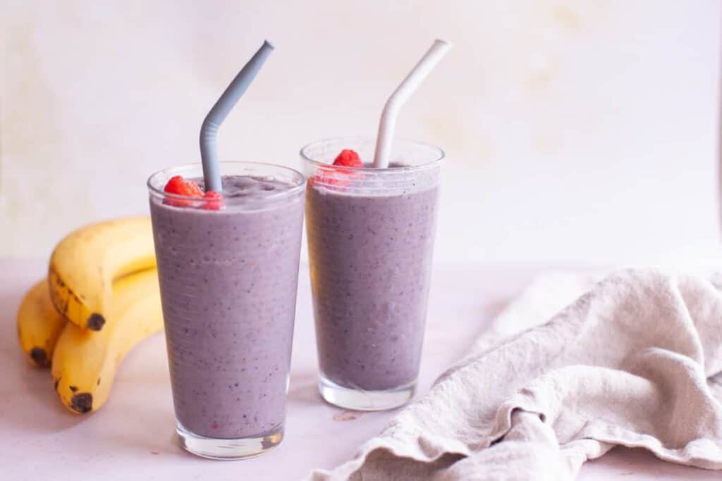 A wide shot of two glasses with straws filled with the smoothie topped with fresh raspberries next to two bananas.