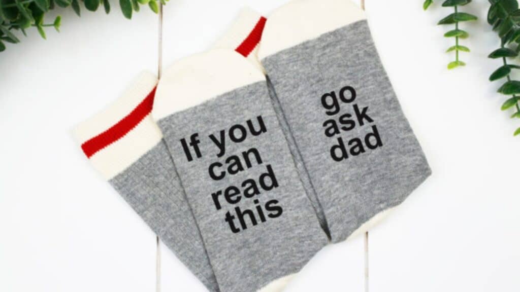 A pair of grey socks with the text "If You Can Read This" and "Go Ask Dad" written on each.