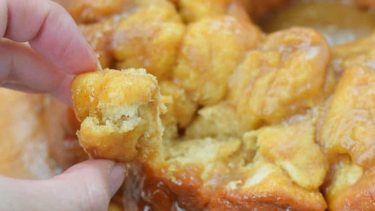 This photo shows a hand reaching out to monkey bread on a white cake platter and pulling off a chunk.