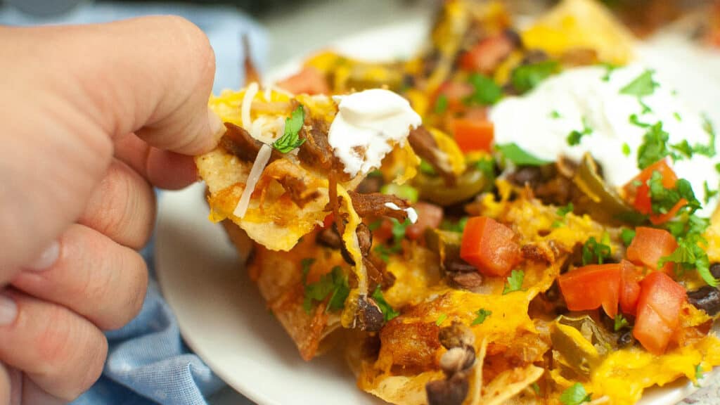 scooping up air fryer nachos topped with cheese, sour cream, tomtao and green onions.