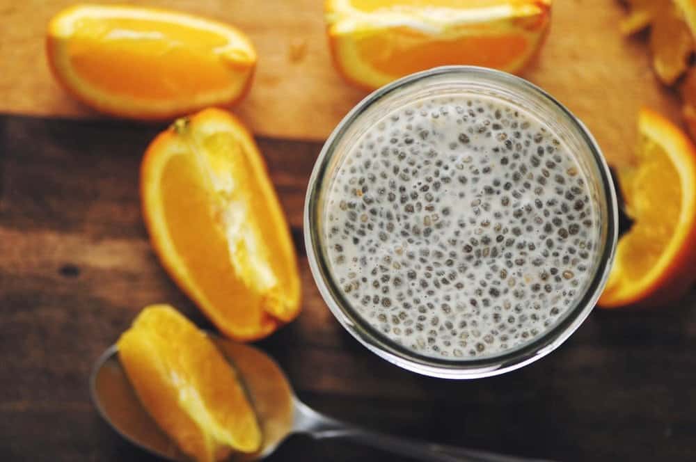 Top view of a clear glass filled with white chia pudding resting on a wooden table next to orange slices.