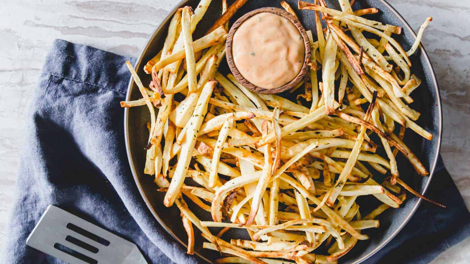 Parsnip fries on a plate with dipping sauce on a blue kitchen towel.