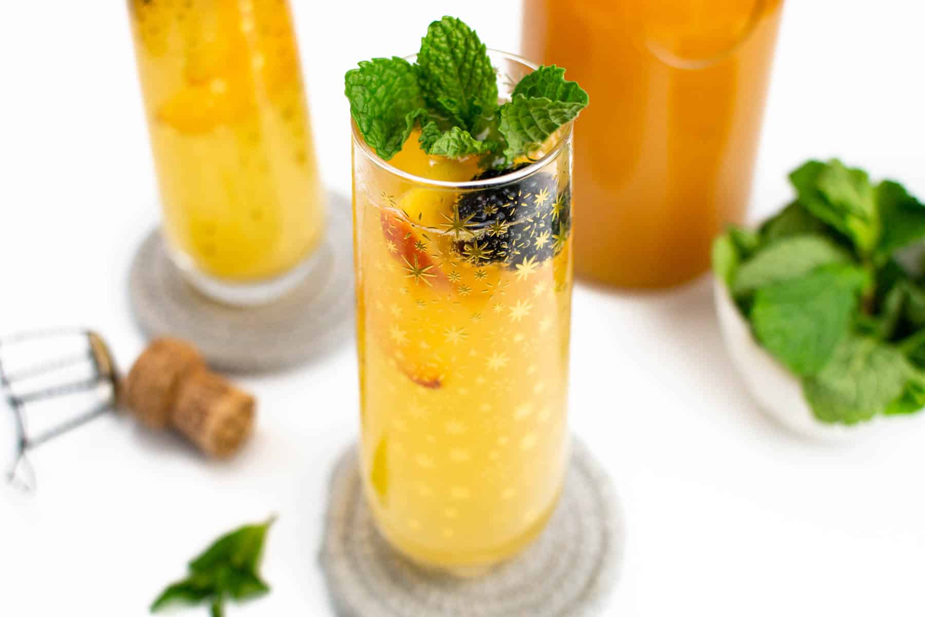 Two peach bellinis sit on woven coasters alongside a bowl of fresh mint, a cork and its cage and a pitcher of peach juice.