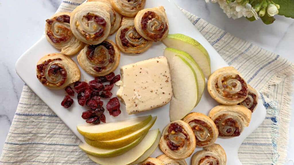Puff pastry appetizer pinwheels on a white board with fresh pears, cranberries and cheese.