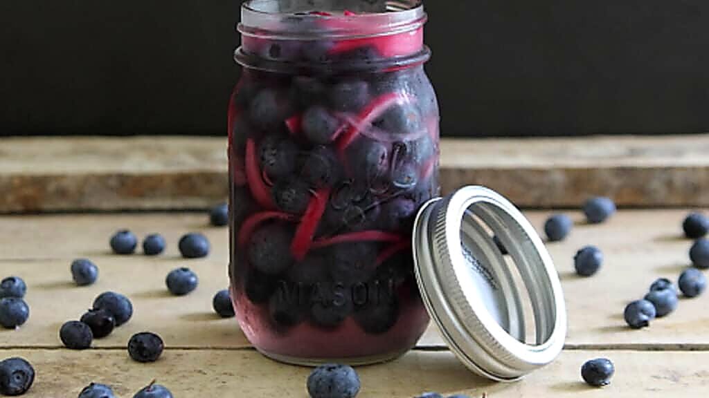 A mason jar filled with a quick pickle of blueberries and red onions sits on a wooden surface, with loose blueberries scattered around and the jar lid off to the side.