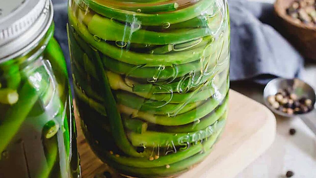 A glass jar filled with quick pickled green beans in brine sits on a wooden surface next to another jar and a spoon with spices.