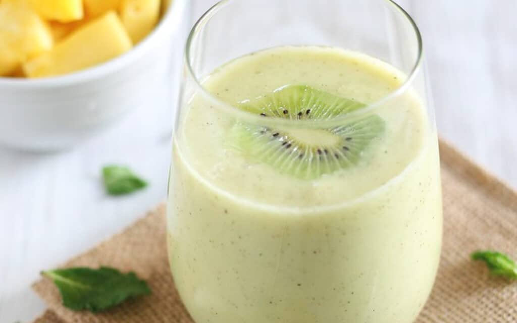 Pineapple kiwi mint smoothie in a glass with kiwi slice garnish.
