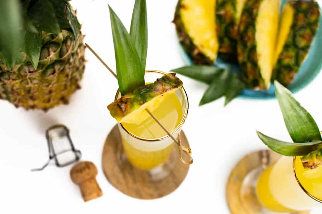 a pineapple, pineapple slices and a champagne stopper next to pineapple cocktails.