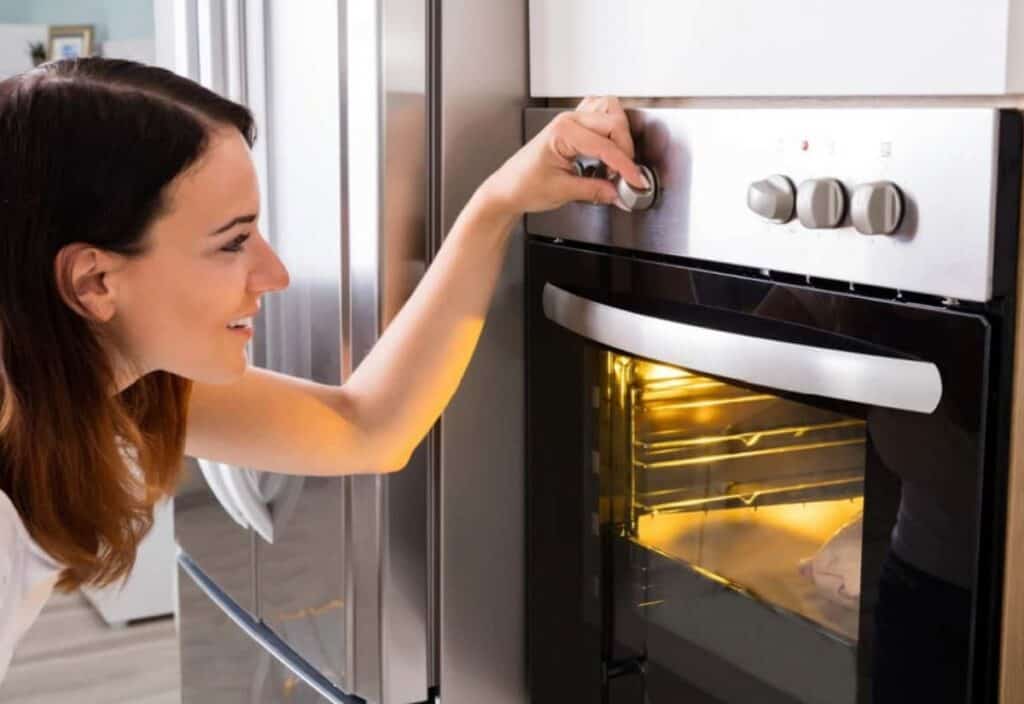 A woman turns the temperature dial on an electric oven with the oven door slightly open and the interior light on.
