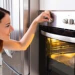 A woman turns the temperature dial on an electric oven with the oven door slightly open and the interior light on.
