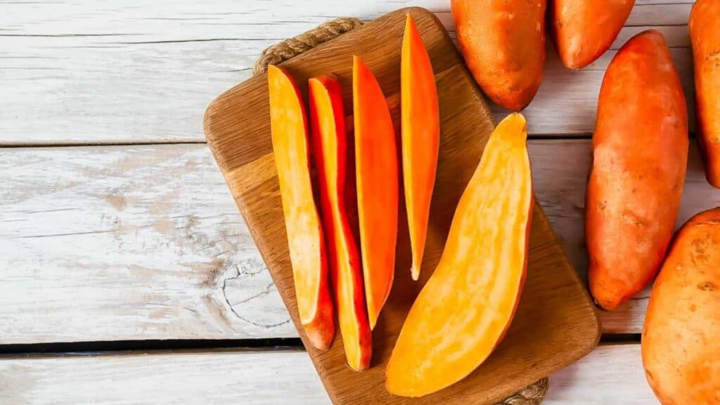 Whole and sliced sweet potatoes on a wooden cutting board, placed on a white wooden surface.