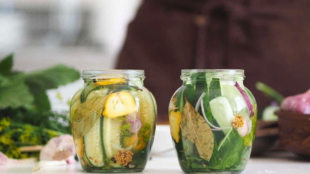 Two glass jars filled with quick pickled cucumbers, herbs, bay leaves, mustard seeds, and yellow vegetables sit on a white surface with greens and onions in the background.