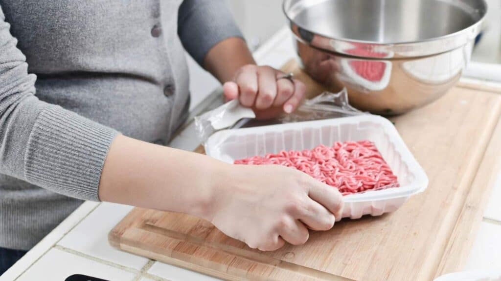 Person opening a plastic tray of raw ground meat on a wooden cutting board, with a metal bowl and knife nearby.