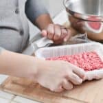 Person opening a plastic tray of raw ground meat on a wooden cutting board, with a metal bowl and knife nearby.