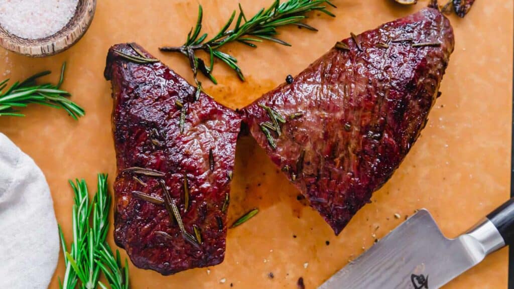 Reverse seared tri tip cut in half on a cutting board with fresh rosemary.