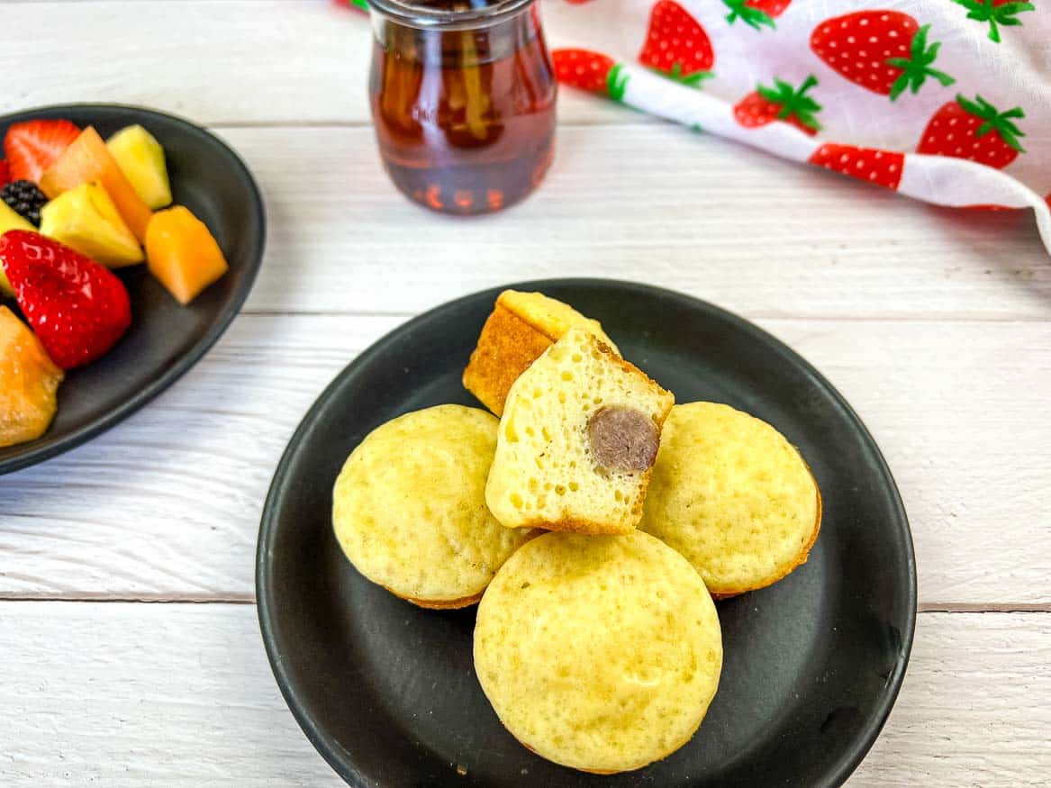 Pancake and sausage bites on a black plate with fruit in the background.