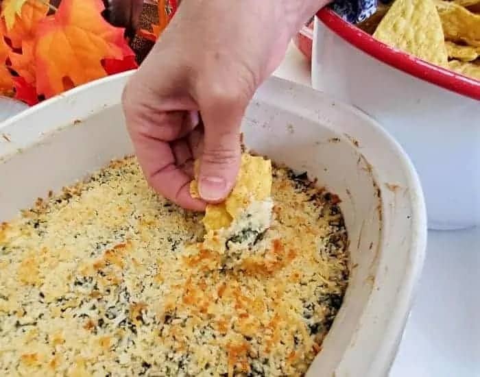 This photo shows a hand scooping out baking spinach artichoke dip from a white casserole dish.