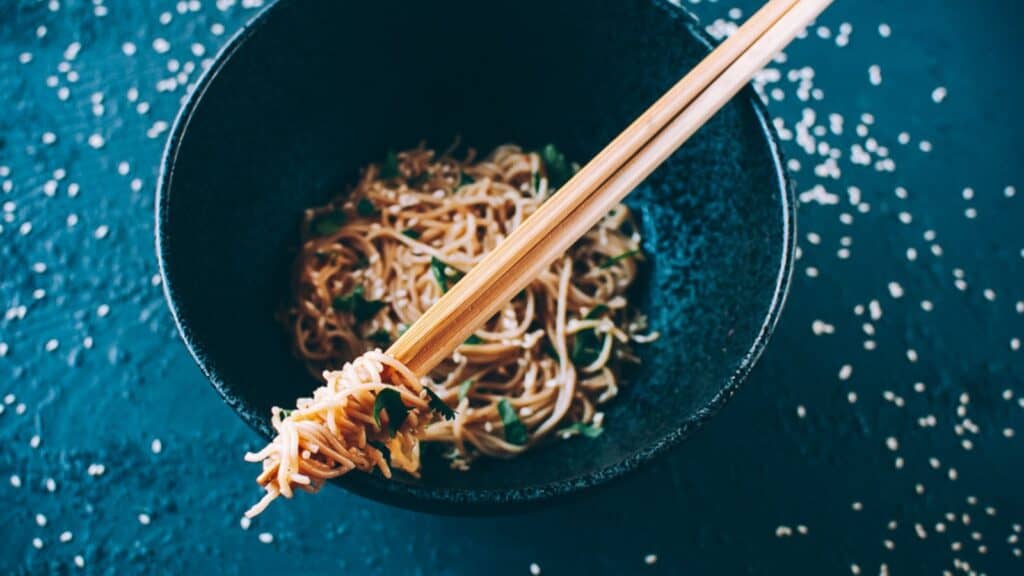 A black ceramic bowl filled with tan noodles and fresh green herbs.