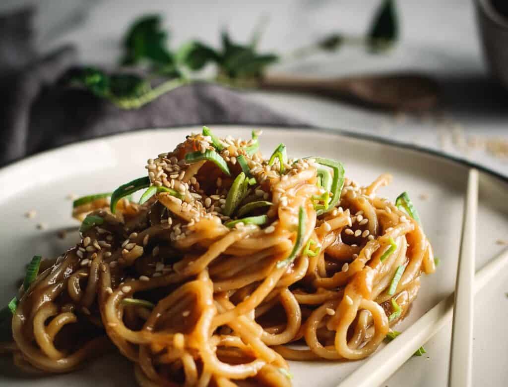 A ceramic plate topped with noodles garnished with sesame seeds and fresh green herbs.