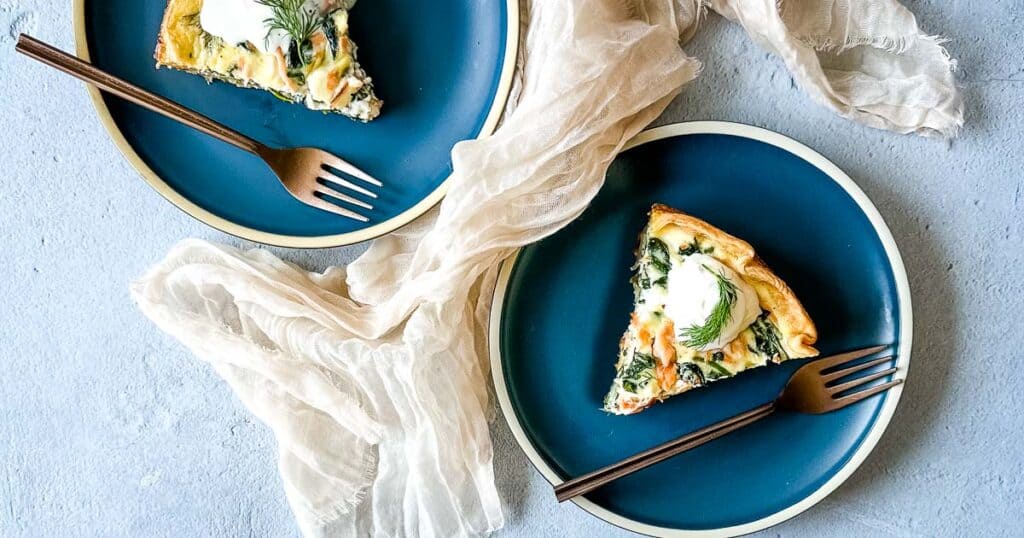 Two blue plates with a slice of smoked salmon and spinach quiche on a gray backdrop.