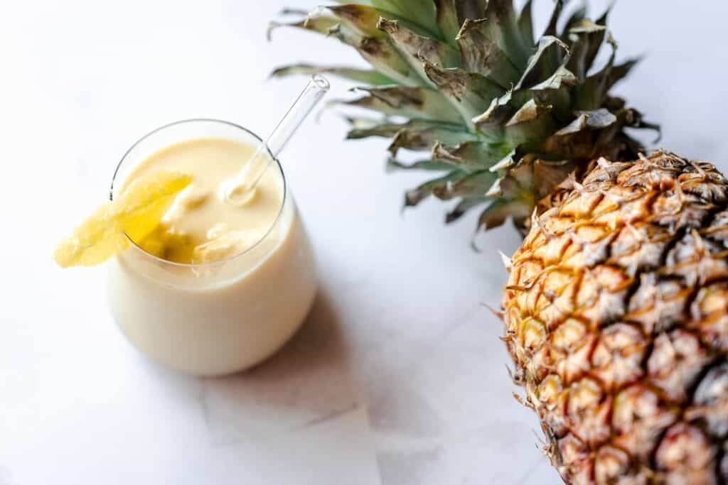 A small clear glass filled with a light yellow smoothie, garnished with a pineapple ring, resting on a gray tabletop next to a large fresh pineapple.