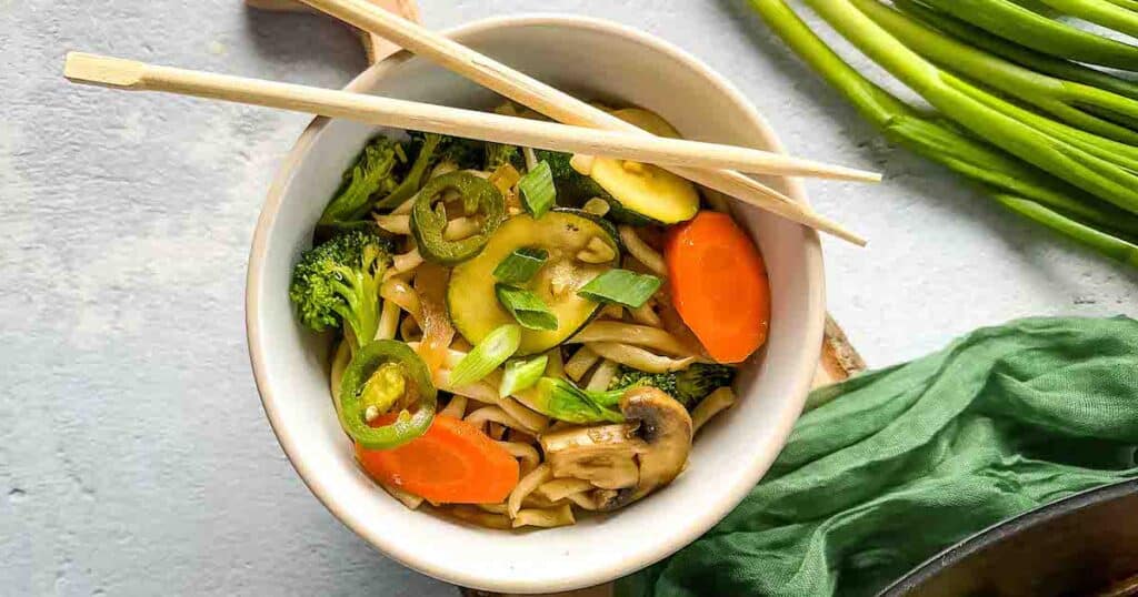 A bowl of vegetable yaki udon is shown with wooden chopsticks, a wok of yaki udon, and scallions in the background.