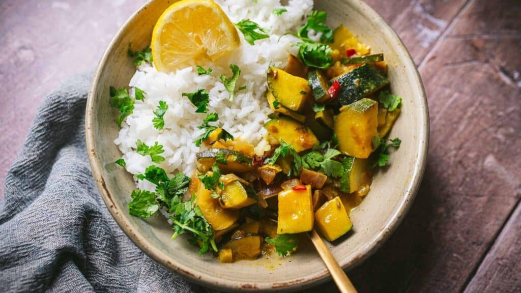 A large tan ceramic bowl of zucchini curry, white rice, fresh herbs and lemon slices.