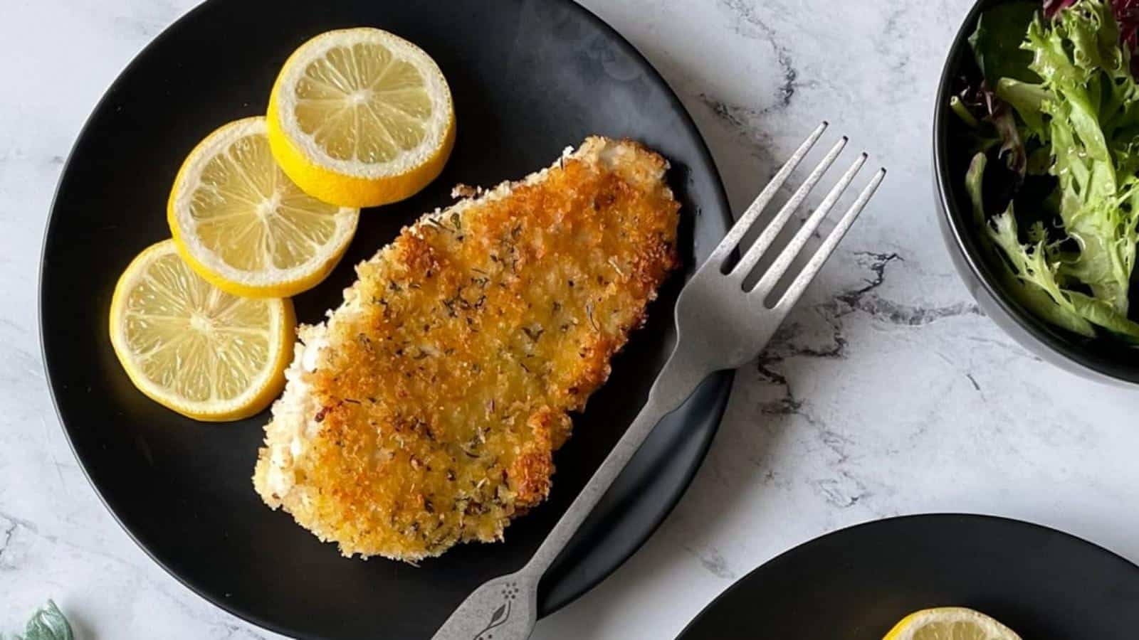 Italian chicken cutlets on a black plate with slices of lemon.