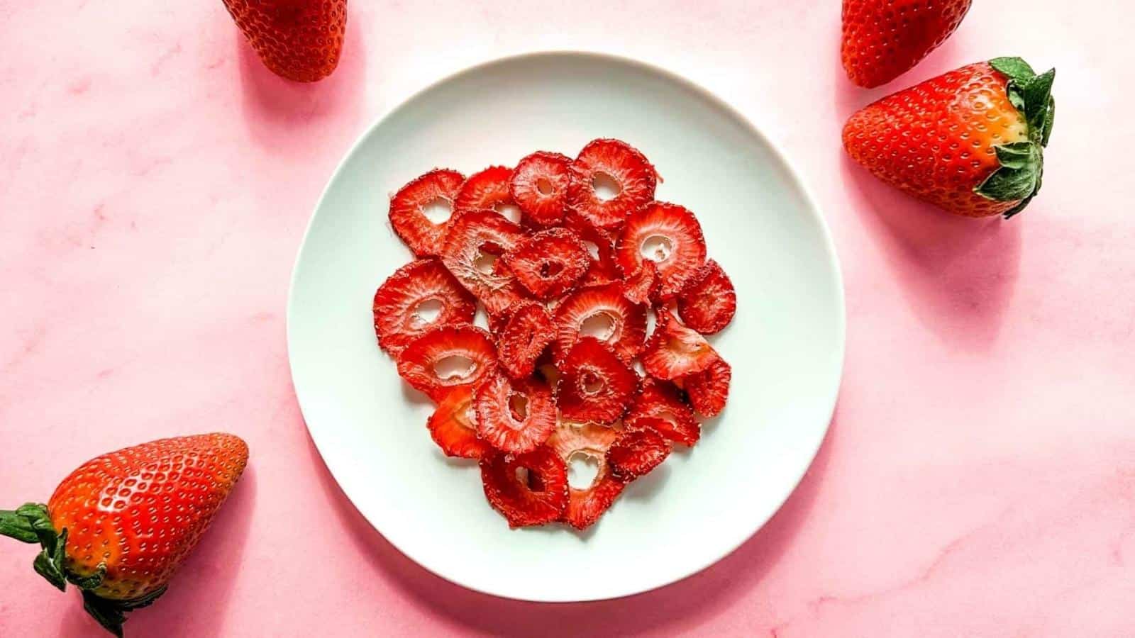 Dehydrated strawberries on a white plate surrounded by fresh strawberries.