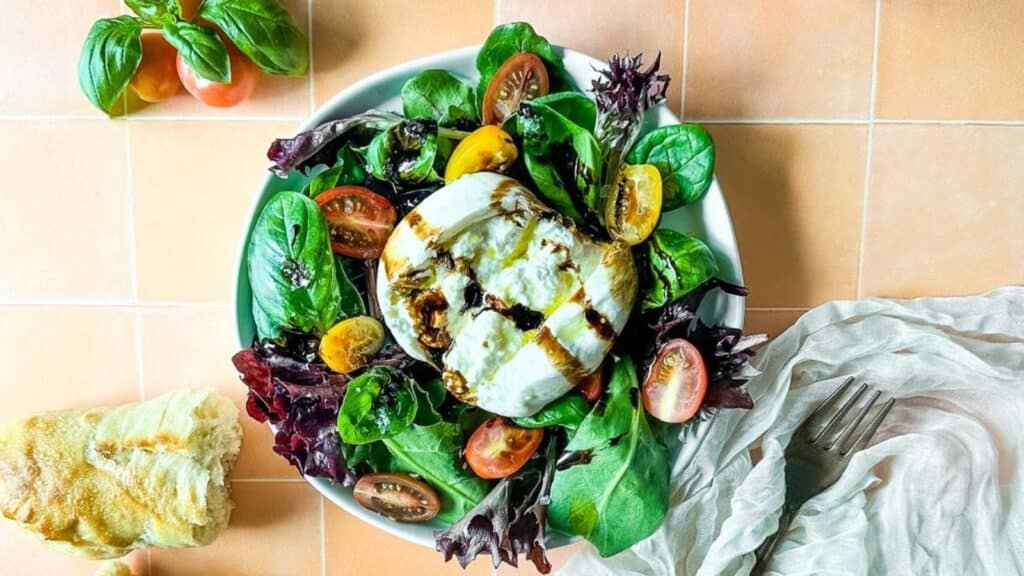 A salad of mixed greens, basil, halved cherry tomatoes, and burrata on a white plate.