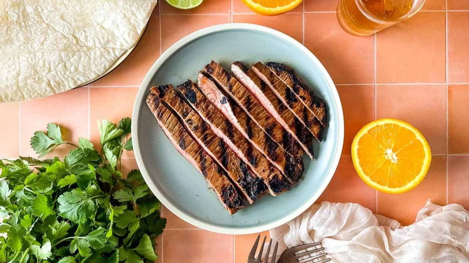 Carne asada on a blue plate surrounded by cilantro, halved oranges, limes, and beer.