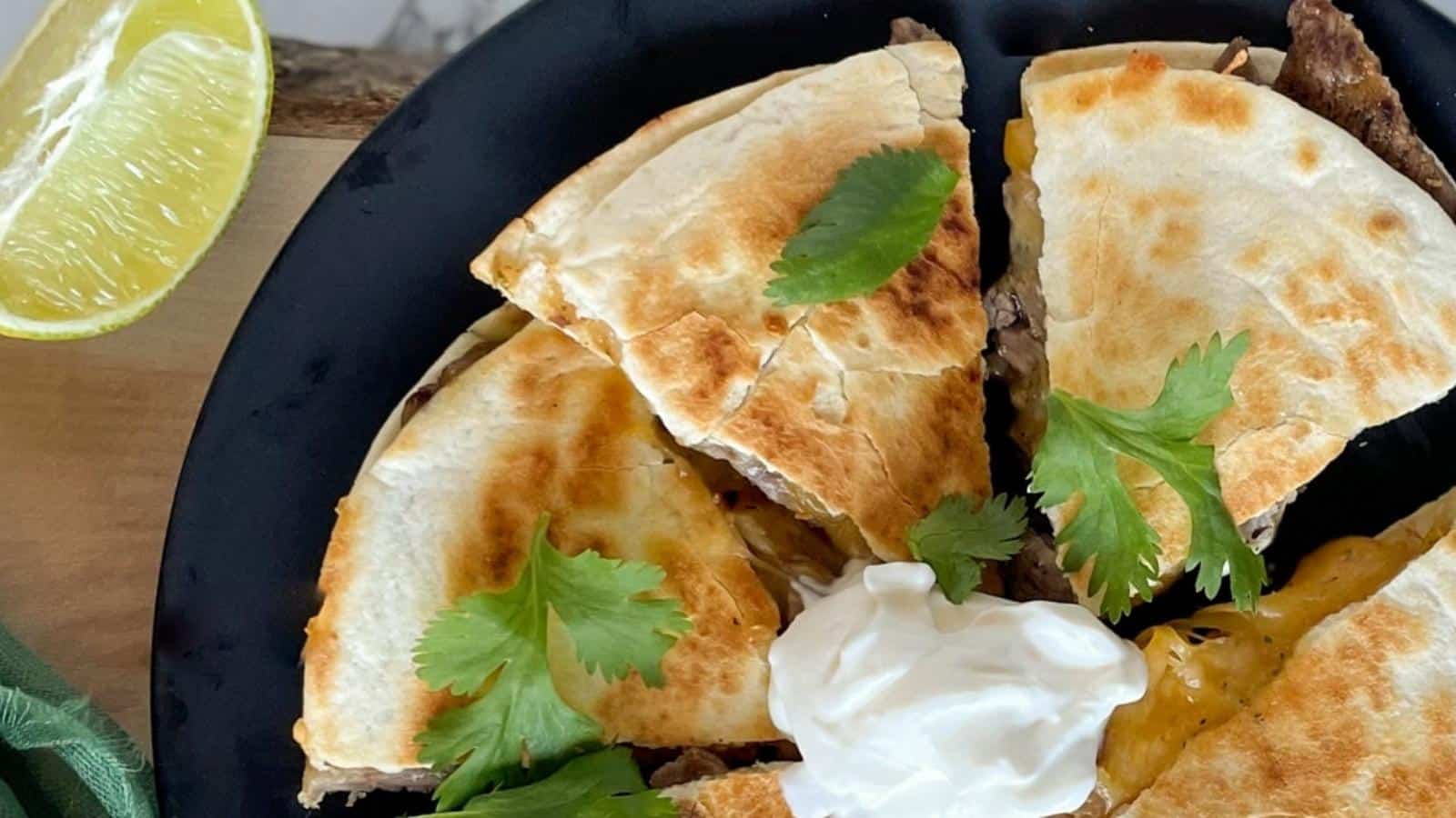 Carne asada quesadilla on a black plate topped with cilantro and sour cream.
