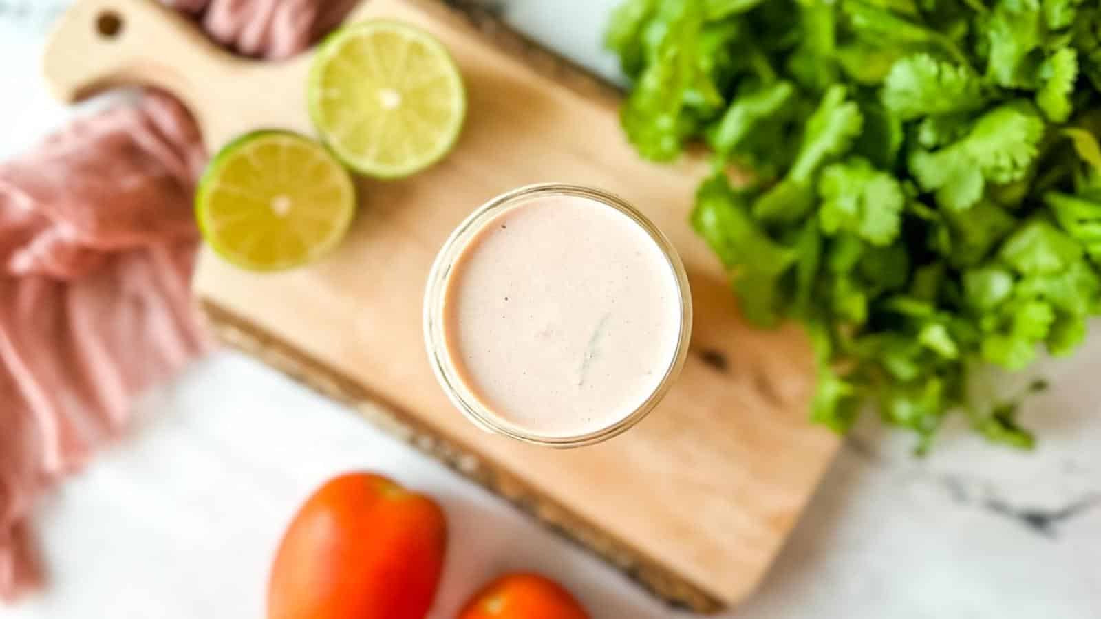 Overhead shot of creamy salsa dressing in a glass jar surrounded by cilantro, fresh tomatoes, and a halved lime.