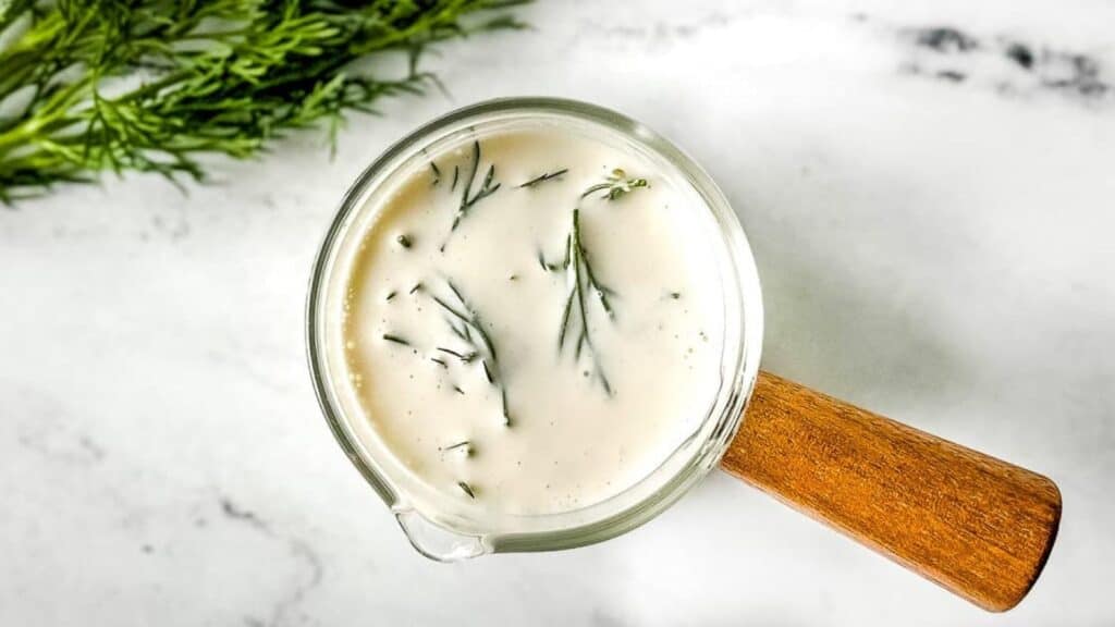 Closeup of dill sauce in a glass container with a wooden handle on a white marble counter.