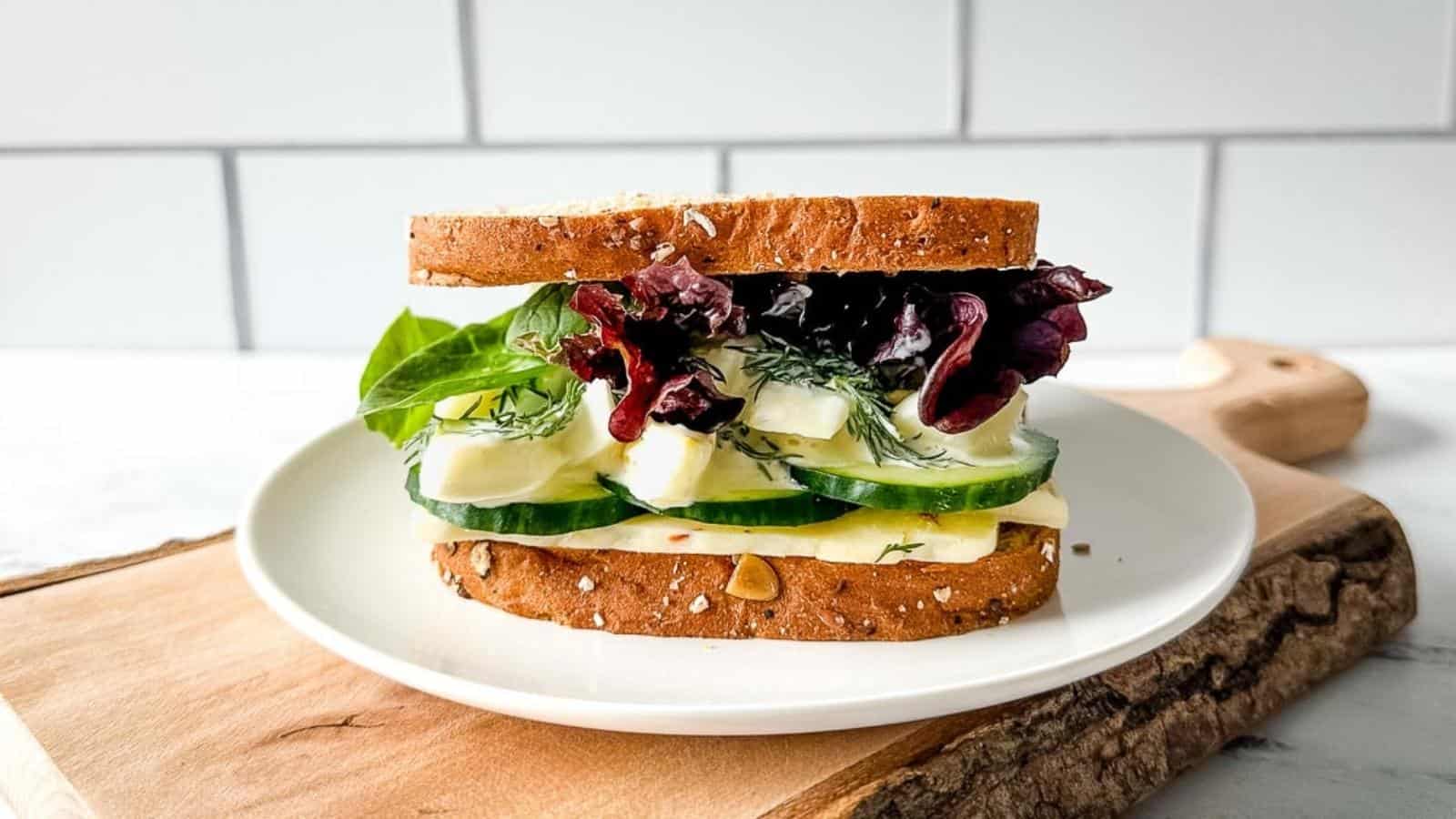 An egg and cucumber sandwich with pepper jack cheese, spring mix, and dill sauce is shown on a white plate over a rustic wooden cutting board.