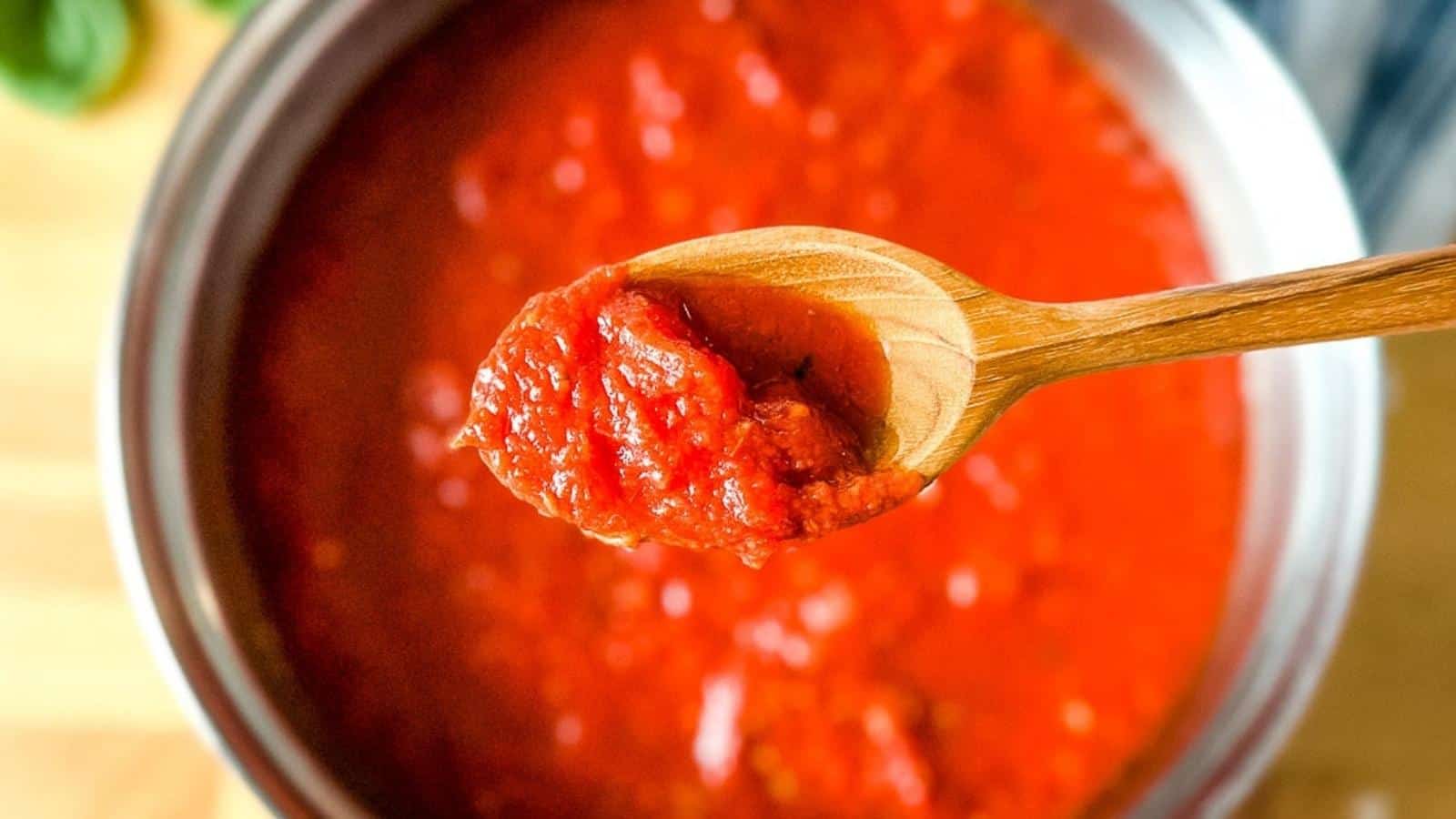 A closeup of hearty marinara sauce in a wooden spoon over a pot of finished marinara sauce.