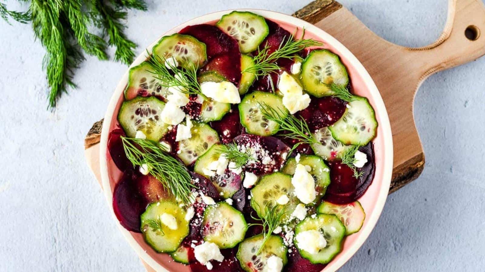 beet cucumber salad is shown on a pink plate with a wood cutting board and dill.