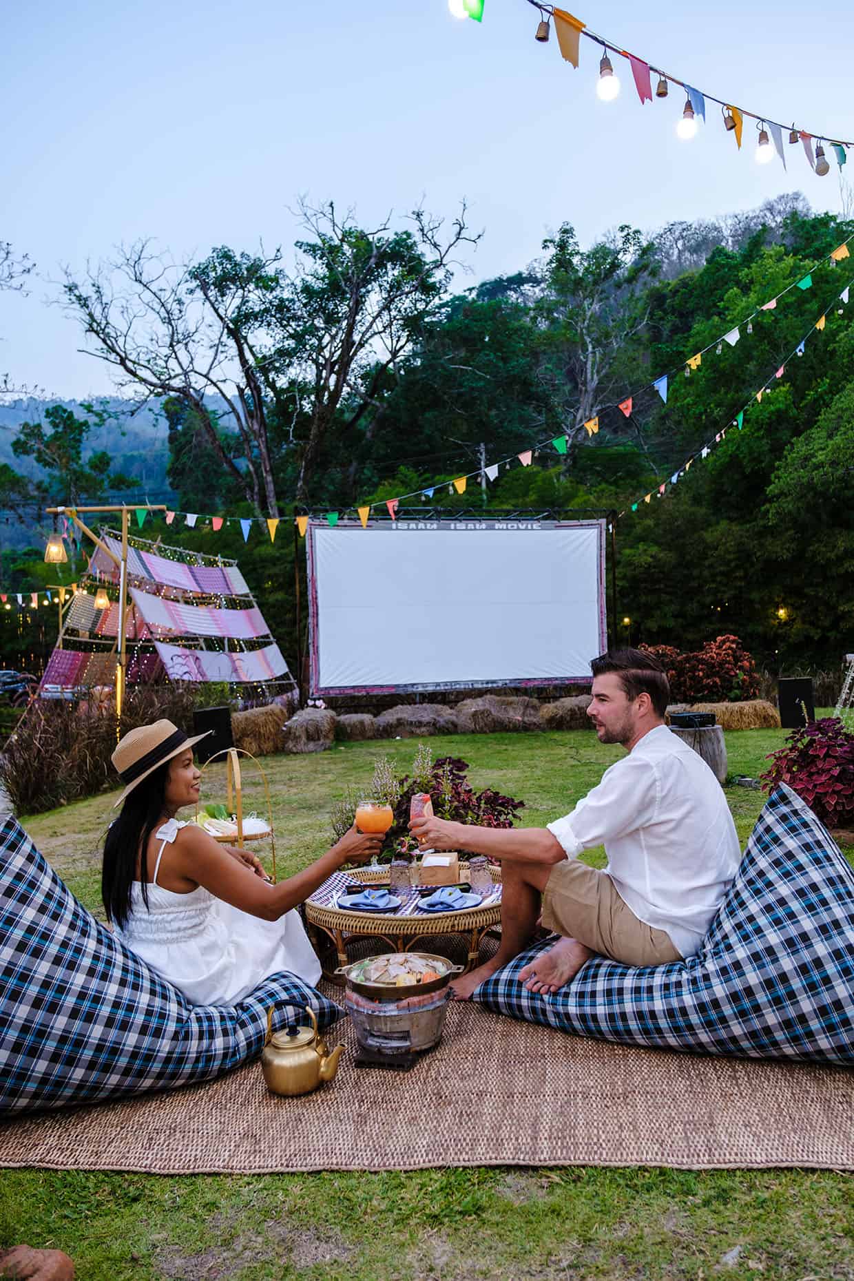 Couple men and women watching a movie in the garden of an outdoor cinema film in a tropical garden with Christmas lights. 