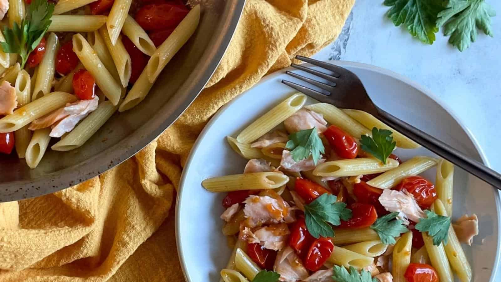 Penne pasta with burst cherry tomatoes, shallots, garlic, and parsley on white plate next to a pan filled with pasta.