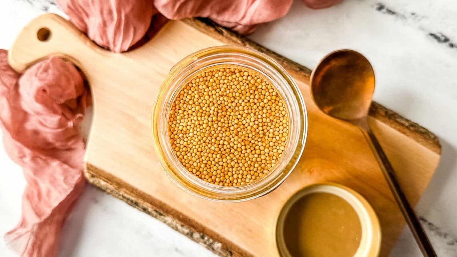 Pickled mustard seeds in a glass jar on a rustic wooden cutting board.