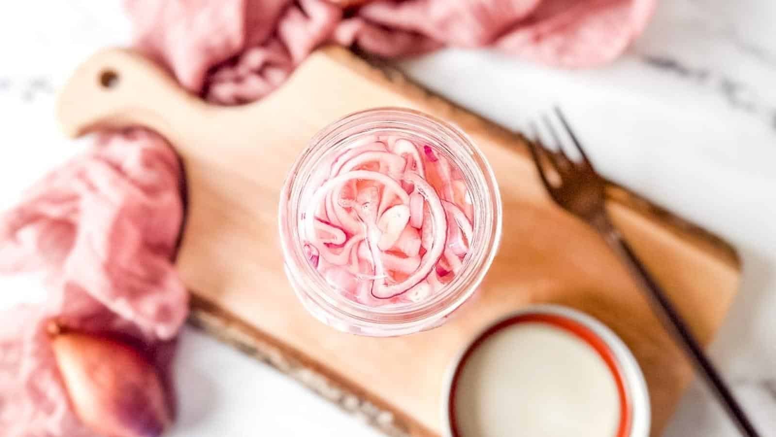 Quick pickled shallots in a glass jar on a rustic wooden cutting board.