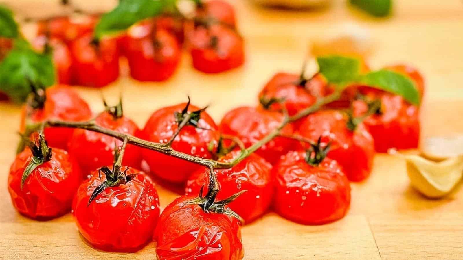 A closeup shot of roasted cherry tomatoes on the vine on a wooden cutting board surrounded by basil and garlic.