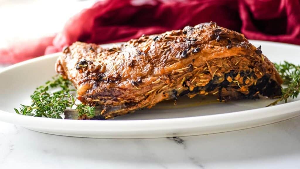 Roasted lamb breast on a white serving platter surrounded by thyme and rosemary.