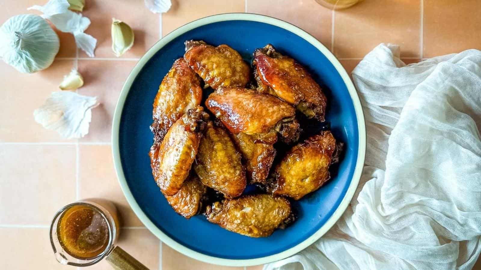 Soy garlic wings on a blue plate surrounded by a white linen, a glass of bear, soy ginger sauce, and cloves of garlic.