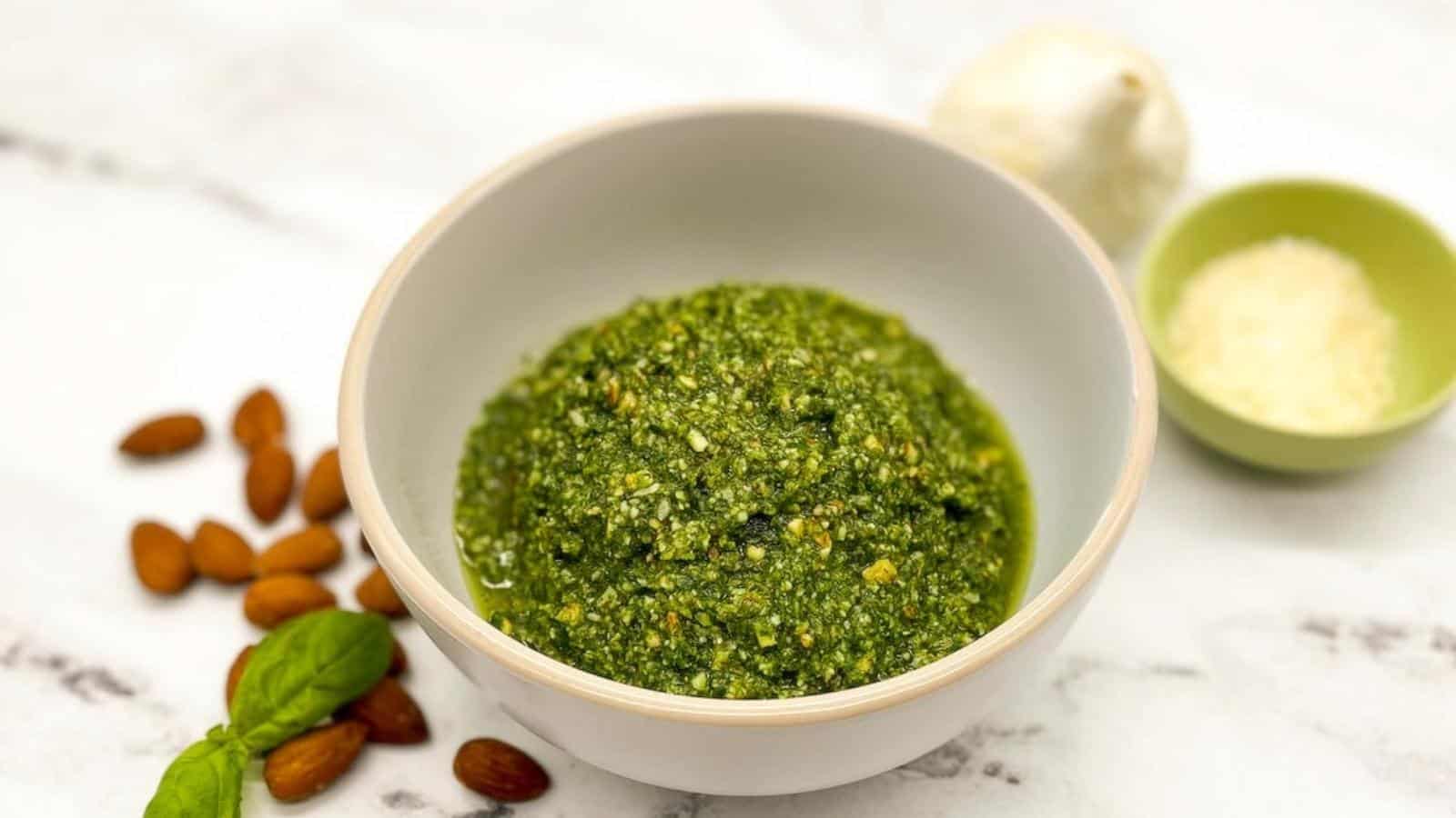 almond pesto in a white bowl surrounded by almonds and basil leaves.