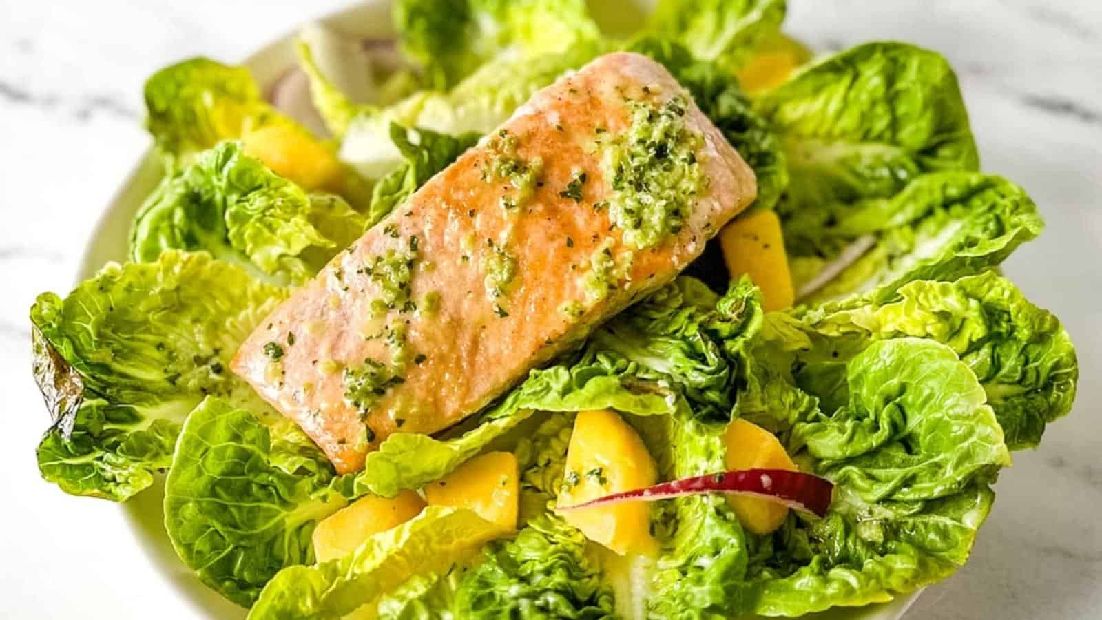 A fillet of salmon sits on a bed of little gem lettuce, mango, red onion, and jalapeno vinaigrette on a white marble counter.