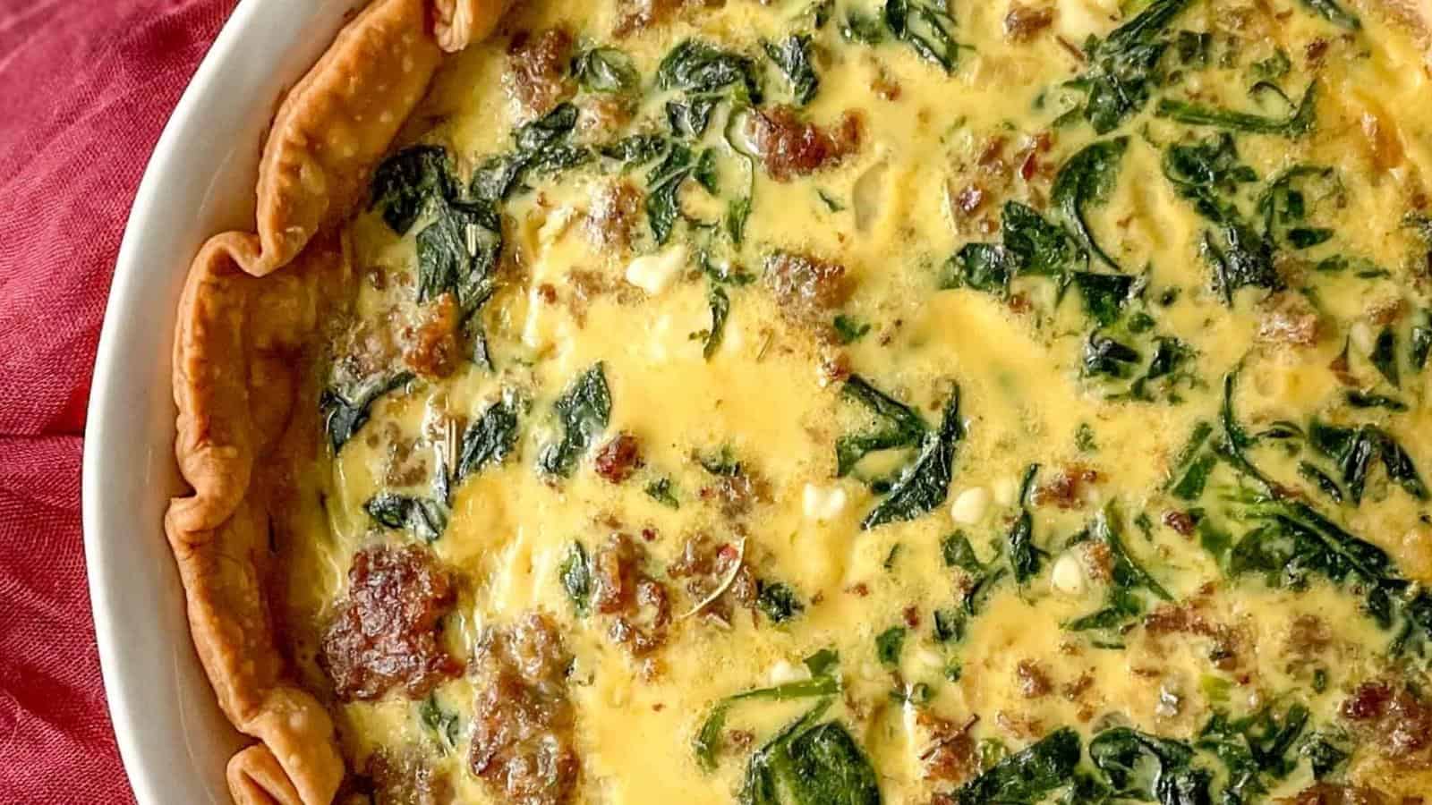 A closeup of a sausage spinach quiche is shown with a red linen.