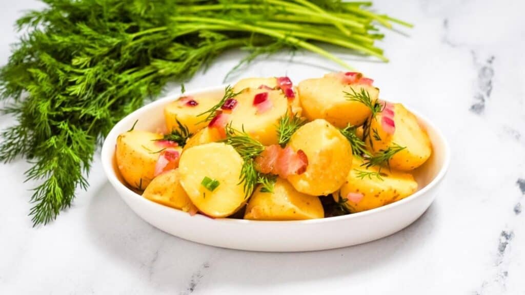 Warm german potato salad in a white dish beside a bunch of dill.